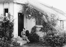 Photograph of Douglas, in her thirties, elegantly dressed, seated on a step outside a cottage which has a low roofline and is surrounded by a profusion of plants.