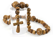 A necklace made out of slightly irregular wooden beads with a small, carved wooden cross in the middle, displayed on top of a transparent plastic cylinder against a white background