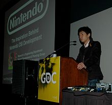 A man stands behind a podium, talking to the audience. Next to him is a projection screen displaying a PowerPoint slide.