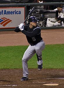 A man in a black baseball jersey and pinstriped baseball pants stands at home plate. He is batting right-handed, holding a black baseball bat over his right shoulder. He is wearing white baseball gloves and a black batting helmet.