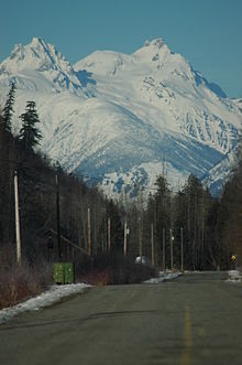 A glaciated mountain rising above trees and a paved road.