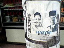 Partially ripped poster of a veiled woman and a man. Chinese text line its vertical edges while Indonesian text is written in the middle.
