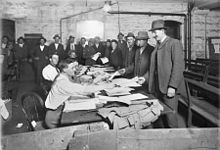 Men in suits and hats file past other men sitting at a long table, handing over paperwork