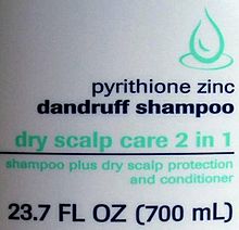 White slightly curved plastic with a green line drawing of a drop, the text "pyrithione zinc dandruff shampoo" in dark blue with "dry scalp care 2 in 1" in green, then a blue line. Underneath it is "shampoo plus dry scalp protection and conditioner" in smaller green type, and finally "23.7 FL OZ (700 mL)" in larger blue type