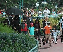 A man in public is dressed in black. He is surrounded by an entourage and members of the public, some of whom are holding cameras. He walks behind three young children, all of whom are wearing facial masks.