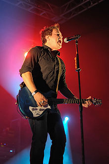 A twenty-something male sings into a standing microphone at a concert venue while playing his electric guitar. Red background lights shine on him.