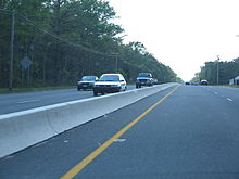 A four lane road in a wooded area with a small concrete barrier in the center. Power lines are on either side of the road.