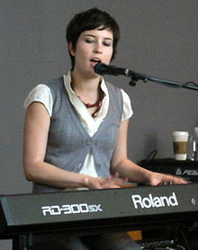 Higgins is seated. She sings into a microphone and plays a keyboard instrument. The lettering RD-300SX and Roland are visible across its front.
