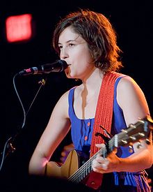 A woman in her twenties with short dark hair plays an acoustic guitar and sings into a microphone, lit by bright stage lights.