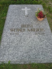 A marbled-grey stone slab, lying flat on the ground, surrounded by grass and weeds, bearing the golden inscription "Oberst Werner Mölders 18.3.1913–22.11.1944" just below the centre of the slab. Above the inscription, there is a small Christian cross and a terracotta bowl with flowers towards the right-hand edge of the slab.