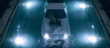 Image of a blond woman. She is sitting over a white sports car, that is inside a pool. She is wet and wears a black dress. The pool is illuminated from above. She is looking up towards the camera.