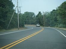 A two lane road lined with power lines making a curve through woodland
