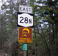 Three signs, from high to low: A rectangular sign that reads "EAST", a New York state highway sign, and a brown-and-yellow trail marker