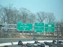 A multilane freeway with a display of three green signs over the road. The left one reads exit 8 east Interstate 287 White Plains Rye three downward arrows, the middle one reads Interstate 87 south Saw Mill Parkway south New York City two downward arrows,and the right one reads exit 8A New York State Route 119 Saw Mill Parkway north Elmsford right lane.