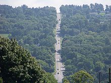 A two-lane highway descends sharply into a valley, where it intersects another roadway at the base of the valley. From there, it ascends the opposite side of the valley and disappears into the background.