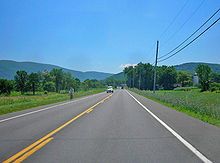 A two-lane highway in a grassy area with a telephone line on the right and mountains in the distance.