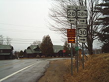 A pair of highways intersect in front of a pair of cabins in a rural area. The highway in the foreground ends here, forcing commuters to turn left or right. A nearby sign assembly indicates that NY 28 west is to the left while NY 28N east and NY 30 north are to the right.