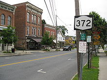 A NY 372 shield attached a post near a telephone pole in the middle of a village
