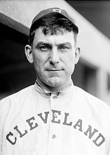 A man with dark hair in a dark baseball cap and a white baseball jersey with "CLEVELAND" on the chest.