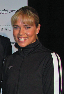 Woman with brown/blonde hair and a fringe, smiling. She is wearing a black Speedo tracksuit, and is standing in front of an advertising wall.