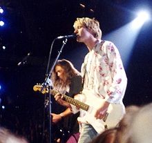 A blonde man wearing a white shirt with flowers plays a white guitar and sings, while in the background another man plays the bass.