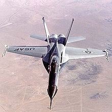 Jet fighter over-flying desert. The jet has black nose and two outward-canted vertical stabilizers between which lay two engines.