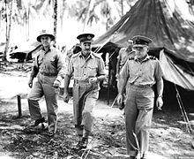 Three men wearing military uniforms walking towards the camera. A tent and palm trees are in the background