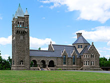 Dark stone-faced building with tall bell tower at left. Arches connect to the main receiving rooms and chapel; roof is a slate blue color.