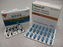 The cardboard packaging of two medications used to treat obesity. Orlistat is shown understanding under the brand name Xenical in a white package with the Roche logo in the bottom right corner ( the Roche name within a hexagon). Sibutramine is below under the brand name Meridia. The package is white on the top and blue on the bottom separated by a measuring tape. The A of the Abbott Laboratories logo is on the bottom half of the package.
