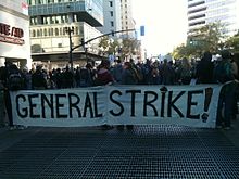 The front of a marching crowd carrying a large banner. The banner reads "General Strike!"