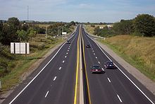 "A photo of a four-lane divided highway on a sunny day. The photo is taken from a position above the centre of the highway, which continues forward into the horizon. Several cars can be seen on the highway. An offramp on either side of the highway merges with it halfway up the picture."
