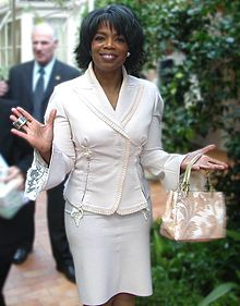 African-American woman, age 50, with short, cropped black hair and wearing a white suit and carrying a white handbag.