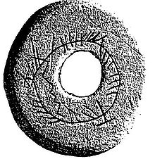 A grainy black-and-white image of a stone disc with a hole in its centre. The surface of the disk has a circle carved into it, which along with the central hole creates a concentric effect. Crude notches, mostly short straight lines, but with some zig-zags, have been carved along and near the circumference of the circle.