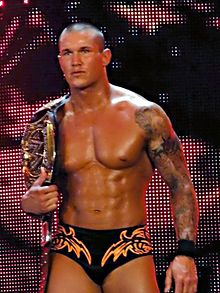 A caucasian male with closely cropped black hair carries a wrestling championship belt over one shoulder. He is wearing short black wrestling tights, with an orange design, and a 'sleeve' tattoo is visible covering one of his arms.