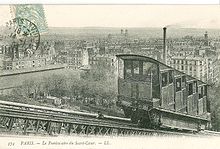 A sepia postcard taken at an angle to the track showing one of the first funicular's cabins. Its stepped compartments are clearly shown. In the background is a panorama of Paris