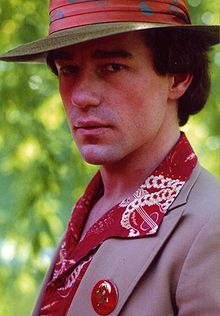 A portrait photo of a man looking side-ways on at the camera with a serious expression on his face. He has a red rimmed hat on, a brown jacket, a gold and red shirt and a button was a man's face on it.