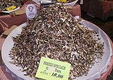 A heaped pile of dried sliced mushrooms on a large plate. A sign near the back of the plate reads "Fungo di Borgotaro I.G.P."; another sign at the front reads "Prezzo speciale di Fiera. 3 Etti 18,00". An electronic scale is partially visible on a table behind the mushrooms