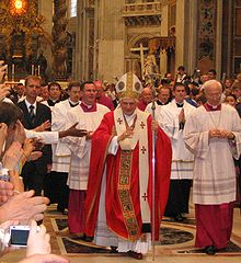  The present Pope, wearing robes of red and white, is walking in procession in St. Peter's, and raising his hand in blessing.
