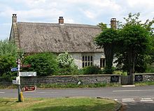 Thatched stone house surrounded by trees. In the foreground a road junction and sign.