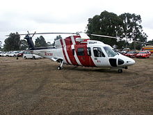 A helicopter painted red and white at rest on grass. Cars are parked in the background.