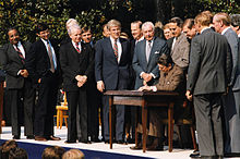 Rangel and about fourteen other men standing around a table, where President Ronald Reagan signs a bill