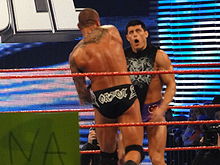 Two dark haired men standing in a wrestling ring with red ropes, one with his back to the camera. The man with his back to the camera is wearing short black wrestling tights with the name 'Orton' written on them in white lettering, and has a tribal design tattoo on his back. He is throwing a punch at the other man, who is facing the camera with a look of surprise, and is wearing short purple wrestling tights and a black top with a white design on it.