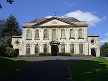 Two storey symmetrical building with two tiers of seven windows. Leading up is a driveway with grass and shrubs on either side.