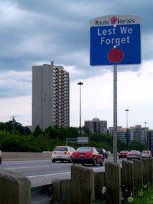 A sign beside a highway with the text Route of Heroes. In the space below is the text Lest we forget, and a red poppy below that.