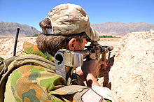 A man wearing a green camouflage uniform and a tan camouflage baseball cap looking through a rifle scope. He is resting his rifle on a dirt wall and steep mountains are visible in the background.