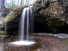A rock outcropping overhanging a small pool of water with a waterfall cascading into the pool.