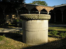 A stout cylindrical column in a courtyard in front of palatial arches of Islamic style