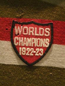 A red shield-shaped cloth crest with the inscription "Worlds Champions 1922–23" sewn onto a gold red and white striped jersey