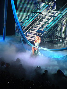 Distant image of a blond woman. She is sitting on a swing hanging from two pieces of fabric. Smoke surrounds her. She is wearing a dress and has her legs crossed. She is holding a microphone and grabbing the swing.