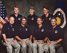 A portrait of six men and one woman, arranged in two rows, four sitting at the front and three standing at the back. They are each wearing tan trousers and a blue polo shirt with a patch and their name on it, and the US and NASA flags are visible in the background.